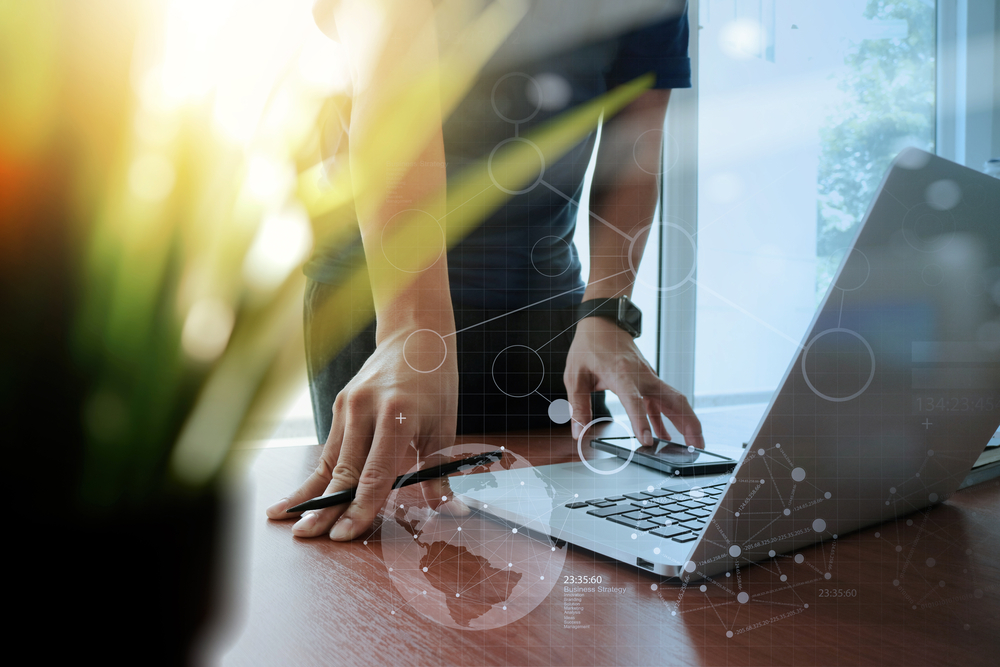 designer hand working laptop with green plant foreground and digital social diagram layers on wooden desk in office